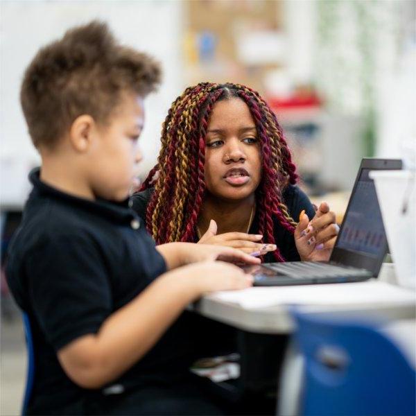 teacher kneels by desk of second grader who is looking at a laptop