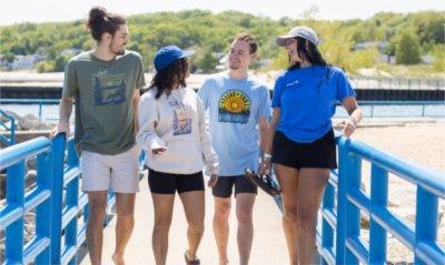 Four students wearing new Lake + Valley clothing items walk along the beach, laughing and talking together.