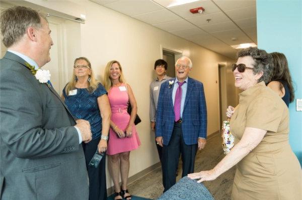 small crowd in hallway of living center, two men wear suits, women in dresses